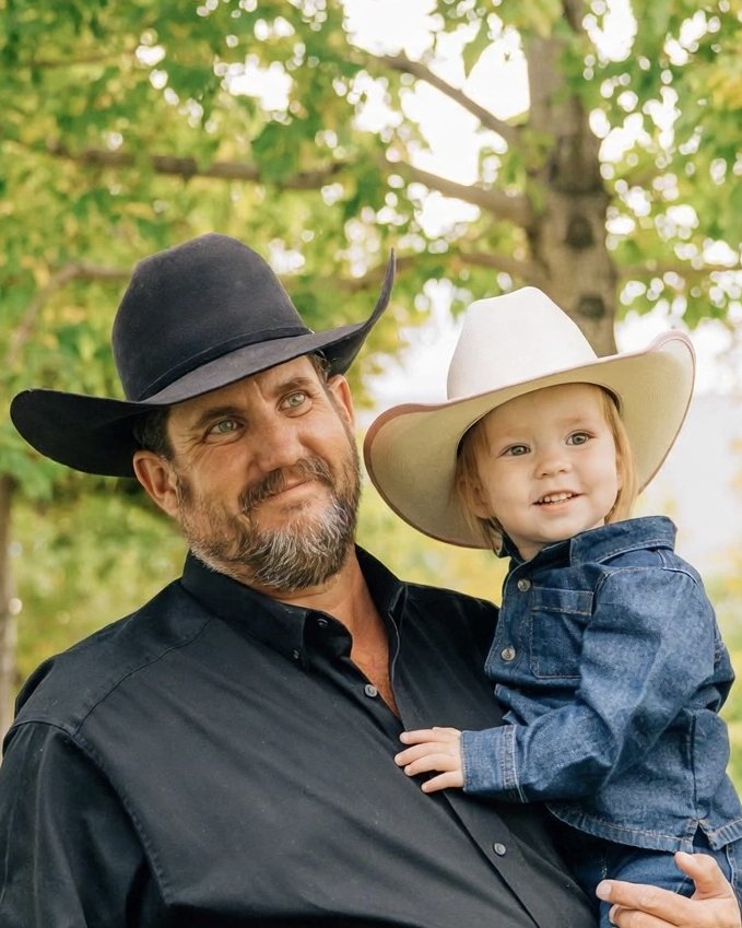 James Henrie holding a child, both wearing cowboy hats, under autumn trees