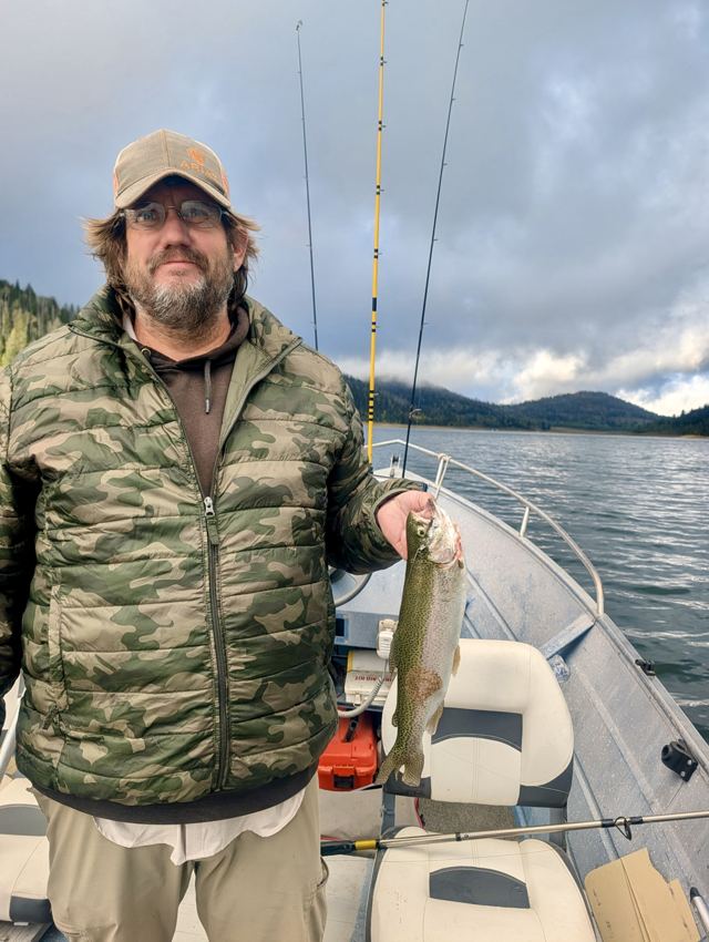Angler in a camouflage jacket holding a rainbow trout on a boat with fishing rods and mountains in the background