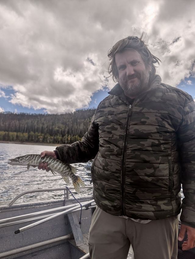 Bearded angler in a camouflage jacket holding a tiger trout on a boat under a cloudy sky with forested hills behind